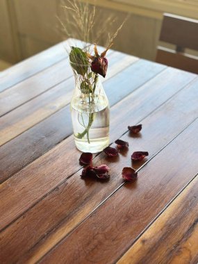 Kurumuş çiçek dalları ve desenli ahşap masaya saçılmış gül yaprakları olan hassas cam bir vazo. Yumuşak ışıklandırma, kompozisyonun sade ve sakin güzelliğini vurguluyor. Huzur ve nostalji uyandırıyor..