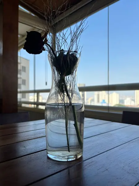 Kurumuş bir gül ve ahşap bir masanın üzerine yerleştirilmiş narin çimen sapları içeren şeffaf cam bir vazo. Arka planda, kentsel görünüm binaları ve berrak gökyüzünü ortaya çıkarıyor, dinginlik ve nostalji uyandırıyor..