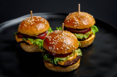Üç sulu mini çizburger, marul, soğan, domates ve erimiş peynirli, susamlı çörekler, siyah arka planda. Fast food, aperatif ya da gurme konsepti için mükemmel. Yüksek kalite fotoğraf