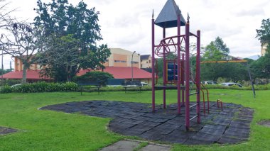 Çerçevesi, salıncağı ve yıpranmış lastik paspasları olan terk edilmiş bir oyun alanı, bina ve ağaçların üzerine kurulmuş..