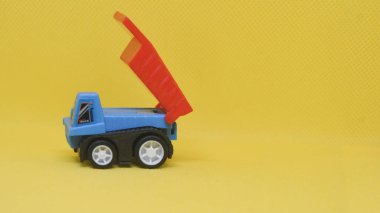Minyatür mavi oyuncak kamyonu, kırmızı konteynırı yukarı eğilmiş, canlı sarı bir arka plana karşı. Çocukluk, oyun ya da inşaat temaları için mükemmel.