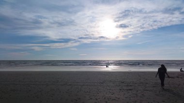 Gün batımında sahilde yürüyen bir insanın silueti, sakin dalgalar ve bulutlu bir gökyüzü, huzuru, yansımayı ve doğanın güzelliğini simgeliyor..