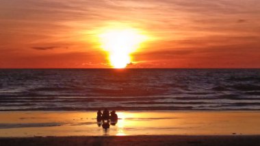 Dostlar plajda toplanıyor, canlı, ateşli bir günbatımına karşı siluet, ıslak kum ve okyanus dalgalarına yansıyan ılık renkler, bağlantıyı, neşeyi ve doğanın güzelliğini simgeliyor..
