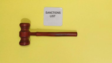 Tahta tokmak, uluslararası ticaret ve finans ile ilgili yasal ve hükümet eylemlerini sembolize eden parlak sarı bir arka planda yer alan yaptırım listesinin yanında yer alıyor. Görüntü bir otorite duygusunu çağrıştırıyor.