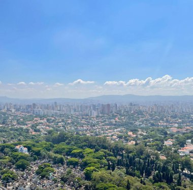 Şehrin çarpıcı panoramik manzarası, yoğun nüfuslu kentsel alanlar ve geniş yeşil alanlar, açık mavi gökyüzünün altında dağınık bulutlarla uyumlu bir kombinasyon gösteriyor. Arka plandaki ağaçlar ve yüksek binalar arasındaki zıtlık