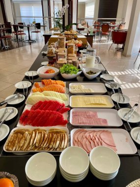 Karpuz, ananas ve papaya gibi taze kesilmiş meyvelerle dolu renkli ve canlı bir kahvaltı büfesinin yanında jambon ve peynir gibi soğuk pirzola. Masa düzgünce düzenlenmiş, sağlıklı bir yemek için davet ediliyor. Arka planda, parlak, modern bir spac