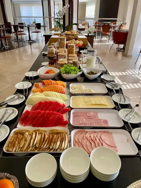Karpuz, ananas ve papaya gibi taze kesilmiş meyvelerle dolu renkli ve canlı bir kahvaltı büfesinin yanında jambon ve peynir gibi soğuk pirzola. Masa düzgünce düzenlenmiş, sağlıklı bir yemek için davet ediliyor. Arka planda, parlak, modern bir spac