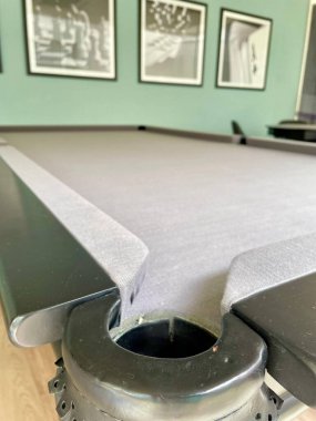 Bir bilardo masasının yanında yakın plan bir resim, üst ve siyah kenarın pürüzsüz gri dokusunu vurguluyor. Arka planda, siyah-beyaz resimlerle dekore edilmiş bir duvar rahat ve modern bir atmosfer yaratıyor..
