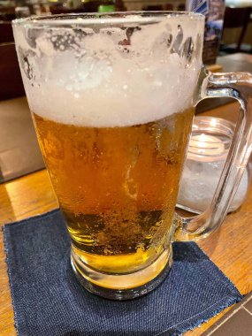 Köpükle kaplı bir bardak soğuk bira koyu mavi bir masa örtüsünün üzerinde duruyor. Altın sıvı ortam ışığını yansıtır, rahat ve rahatlatıcı bir atmosfer yaratır, bir bar veya restoranda gevşemek için mükemmeldir..