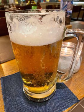 Üstünde köpük olan soğuk bir bardak bira, sıcak bir ortamda koyu mavi bir masa örtüsünün üzerinde duruyor. Yumuşak bir ışık sahneyi aydınlatırken, bir mum romantik bir dokunuş ekliyor. Altın bira ışığı yansıtır. Kadeh kaldırır ve dinlenir..