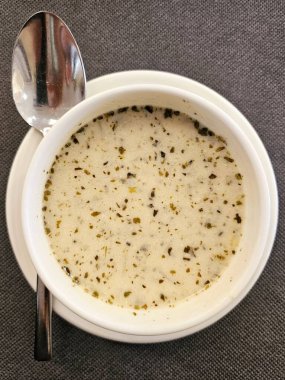 Yeşil naneli yoğurt çorbası, beyaz bir kasede servis ediliyor. Çorbanın pürüzsüz bir dokusu ve görünen bitkiler var. Baharat ya da garnitür içeriyor. Eşleşen bir fincan tabağı ve metal bir kaşık gri bir kumaş üzerinde sola yerleştiriliyor..