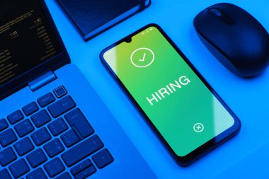 Ofis masasında bilgisayar faresi ve dizüstü bilgisayarlı modern akıllı telefon ekranda işe alma metnini gösteriyor