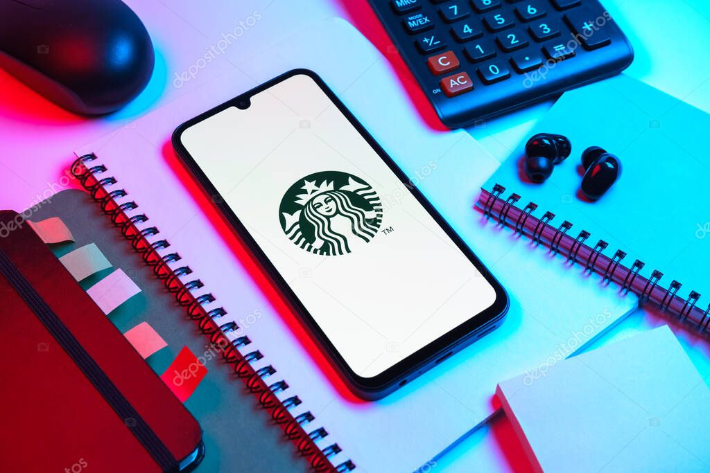 Prague, Czechia - 08 18 2025: An arrangement of office supplies with a mobile phone in the center displaying Starbucks logo. Starbucks is a global coffeehouse chain serving beverages and food.