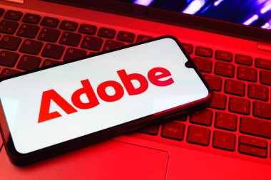 Prag, Çek Cumhuriyeti - 08 01 2025: Dizüstü bilgisayarın üzerinde duran bir cep telefonu Adobe logosunu gösterir. Adobe yaratıcı araçlar ve hizmetler sunan bir yazılım şirketidir..