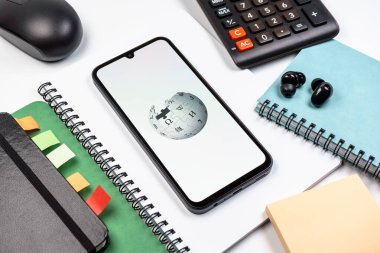 Prag, Çek Cumhuriyeti - 08 18 2025: Merkezinde Wikipedia logosu bulunan bir cep telefonu ile ofis malzemeleri düzenlemesi. Vikipedi çevrimiçi bir ansiklopedidir..