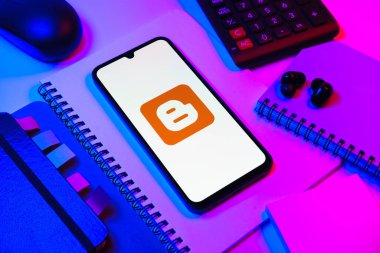 Prag, Çek Cumhuriyeti - 08 18 2025: Blog logosunu gösteren bir cep telefonuyla ofis malzemeleri düzenlemesi. Blogger blog oluşturma ve yayınlama için bir platformdur.