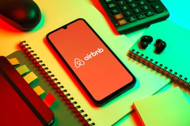 Prag, Çek Cumhuriyeti - 08 18 2025: Merkezde Airbnb logosu sergilenen bir cep telefonu ile ofis malzemeleri düzenlemesi. Airbnb dünya çapında kısa vadeli konaklama için bir platformdur..