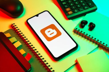 Prag, Çek Cumhuriyeti - 08 18 2025: Blog logosunu gösteren bir cep telefonuyla ofis malzemeleri düzenlemesi. Blogger blog oluşturma ve yayınlama için bir platformdur.
