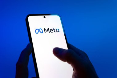 Prague, Czechia - 08 12 2025: Hands on a blue background are holding a mobile phone displaying the Meta logo. Meta is a company offering social platforms like Facebook and Instagram.