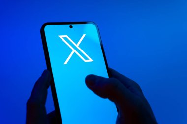Prague, Czechia - 08 12 2025: Hands on a blue background are holding a mobile phone displaying the X logo. X is a platform for sharing short posts and updates.