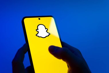 Prague, Czechia - 08 12 2025: Hands on a blue background are holding a mobile phone displaying the Snapchat logo. Snapchat is an app for sending photos, videos, and temporary messages.