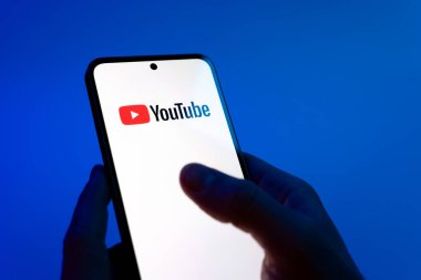 Prague, Czechia - 08 12 2025: Hands on a blue background are holding a mobile phone displaying the Youtube logo. Youtube is a platform to watch and share videos worldwide.