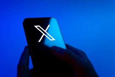 Prague, Czechia - 08 12 2025: Hands on a blue background are holding a mobile phone displaying the X logo. X is a platform for sharing short posts and updates.