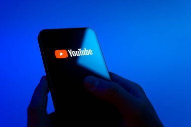 Prague, Czechia - 08 12 2025: Hands on a blue background are holding a mobile phone displaying the Youtube logo. Youtube is a platform to watch and share videos worldwide.
