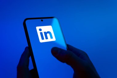 Prague, Czechia - 08 12 2025: Hands on a blue background are holding a mobile phone displaying the LinkedIn logo. LinkedIn is a professional network connecting people and businesses.