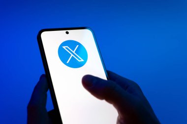 Prague, Czechia - 08 12 2025: Hands on a blue background are holding a mobile phone displaying the X logo. X is a platform for sharing short posts and updates.