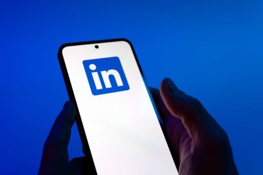 Prague, Czechia - 08 12 2025: Hands on a blue background are holding a mobile phone displaying the LinkedIn logo. LinkedIn is a professional network connecting people and businesses.