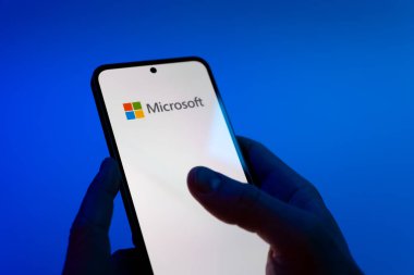 Prague, Czechia - 08 12 2025: Hands on a blue background are holding a mobile phone displaying the Microsoft logo. Microsoft is a technology company offering software, hardware, and services.