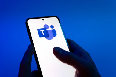 Prague, Czechia - 08 12 2025: Hands on a blue background are holding a mobile phone displaying the Teams logo. Teams is a platform for team collaboration, meetings, and communication.