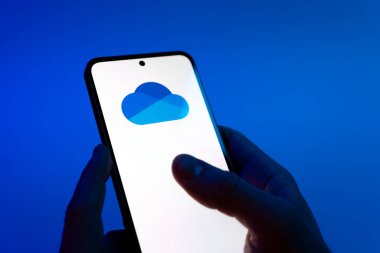 Prague, Czechia - 08 12 2025: Hands on a blue background are holding a mobile phone displaying the OneDrive logo. OneDrive is a cloud storage platform for saving and sharing files.