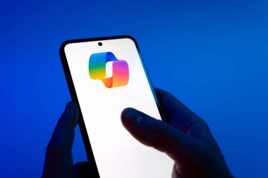 Prague, Czechia - 08 12 2025: Hands on a blue background are holding a mobile phone displaying the Copilot logo. Copilot is an AI assistant integrated into software for productivity help.