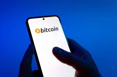 Prague, Czechia - 08 12 2025: Hands on a blue background are holding a mobile phone displaying the Bitcoin logo. Bitcoin is a digital cryptocurrency for decentralized transactions.