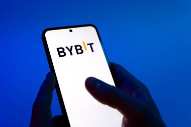 Prague, Czechia - 08 12 2025: Hands on a blue background are holding a mobile phone displaying the Bybit logo. Bybit is a platform for trading cryptocurrencies and derivatives.
