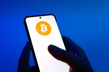 Prague, Czechia - 08 12 2025: Hands on a blue background are holding a mobile phone displaying the Bitcoin logo. Bitcoin is a digital cryptocurrency for decentralized transactions.