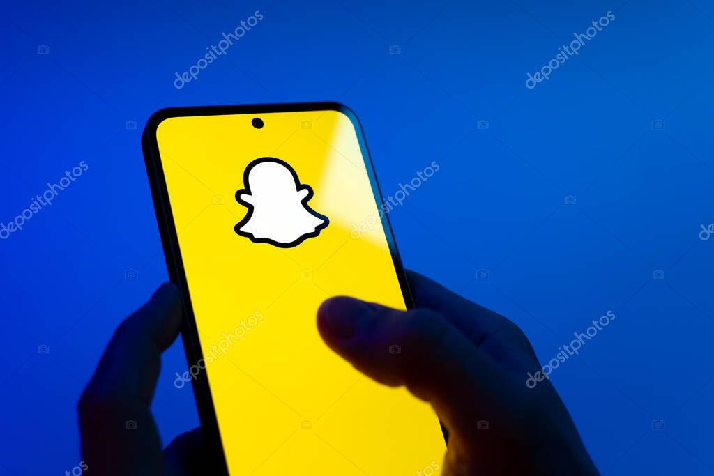 Prague, Czechia - 08 12 2025: Hands on a blue background are holding a mobile phone displaying the Snapchat logo. Snapchat is an app for sending photos, videos, and temporary messages.
