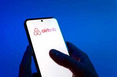 Prague, Czechia - 08 12 2025: Hands on a blue background are holding a mobile phone displaying the Airbnb logo. Airbnb is a platform for booking short-term accommodations worldwide.