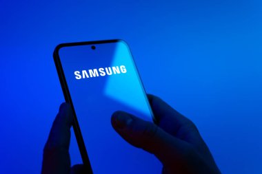 Prague, Czechia - 08 12 2025: Hands on a blue background are holding a mobile phone displaying the Samsung logo. Samsung is a technology company making electronics and devices.