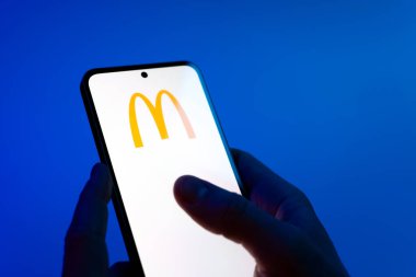Prague, Czechia - 08 12 2025: Hands on a blue background are holding a mobile phone displaying the McDonalds logo. McDonalds is a fast-food chain serving burgers, fries, and more.