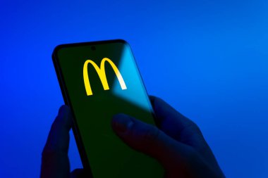 Prague, Czechia - 08 12 2025: Hands on a blue background are holding a mobile phone displaying the McDonalds logo. McDonalds is a fast-food chain serving burgers, fries, and more.