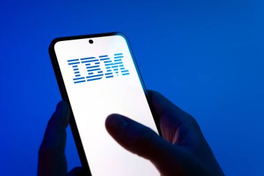 Prag, Çek Cumhuriyeti - 08 12 2025: Mavi arkaplandaki eller IBM logosunu gösteren bir cep telefonu tutuyor. IBM yazılım, donanım ve danışmanlık sunan bir teknoloji şirketidir..