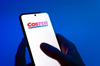 Prag, Çek Cumhuriyeti - 08 12 2025: Mavi arkaplandaki eller Costco logosunu gösteren bir cep telefonu tutuyor. Costco toptan satış yapan bir perakendecidir. Üyelere toptan ürün satar..