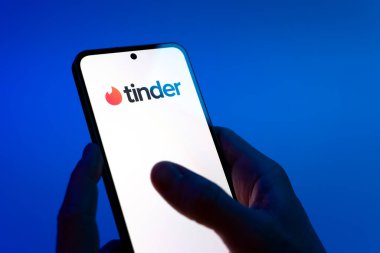 Prag, Çek Cumhuriyeti - 08 12 2025: Mavi arkaplandaki eller Tinder logosunu gösteren bir cep telefonu tutuyor. Tinder insanları eşleştirmek ve bağlamak için bir randevu uygulaması.