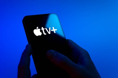 Prag, Çek Cumhuriyeti - 08 12 2025: Mavi arkaplandaki eller Apple TV + logosunu gösteren bir cep telefonu tutuyor. Apple TV +, filmler ve TV programları için bir yayın platformudur..