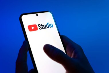 Prag, Çek Cumhuriyeti - 08 12 2025: Eller mavi arkaplanda Youtube Stüdyo logosunu gösteren bir cep telefonu tutuyor. Youtube Studio YouTube kanallarını yönetmek ve analiz etmek için bir platformdur.