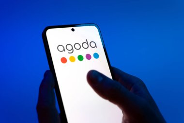 Prag, Çek Cumhuriyeti - 08 12 2025: Mavi arkaplandaki eller Agoda logosunu gösteren bir cep telefonu tutuyor. Agoda, otelleri ve seyahat yerlerini ayırtmak için bir platform..
