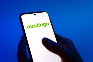 Prag, Çek Cumhuriyeti - 08 12 2025: Mavi arkaplandaki eller Duolingo logosunu gösteren bir cep telefonu tutuyor. Duolingo interaktif olarak dil öğrenmek için bir platformdur..